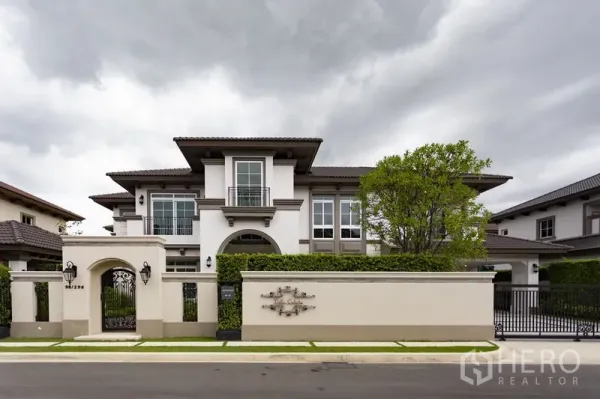 独立式住宅 出租在Mueang Samut Prakan, Samut Prakan - 两层独栋别墅正立面，围墙与绿篱庭院，天空多云。