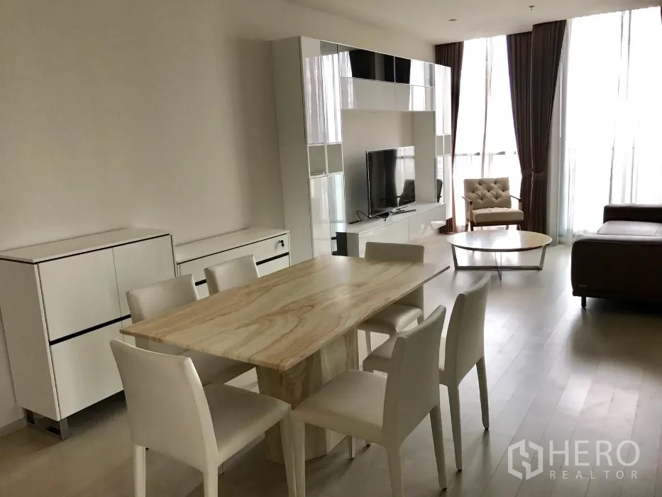 Condominium for rent or sale in Pathum Wan, Bangkok - Bright living and dining area with TV wall unit, sofa, armchair, and floor-to-ceiling windows with curtains.