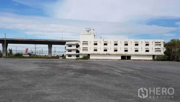 Shophouse for rent in Bang Bo, Samut Prakan - Panoramic view of a large asphalt lot in front of a white building with an expressway nearby.