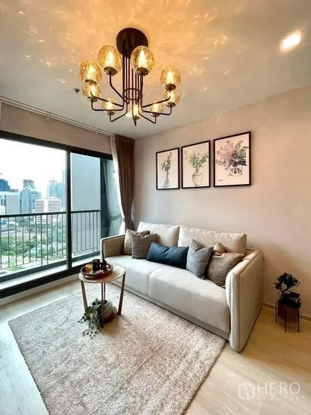 Condominium for rent in Pathum Wan, Bangkok - Bright living room with chandelier, sofa, floral artwork and balcony city skyline view.