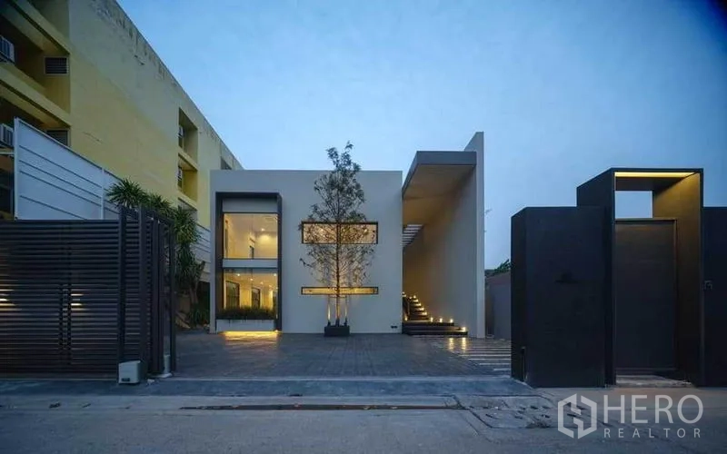 Home Office for sale in Wang Thonglang, Bangkok - Street-facing modern facade with gated entry and wide paved parking court at twilight.