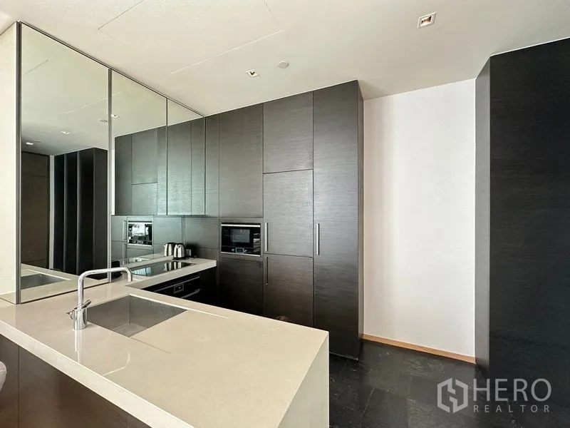 Condominium for rent in Pathum Wan, Bangkok - Modern kitchen with white countertop, mirrored backsplash and dark built-in cabinetry.