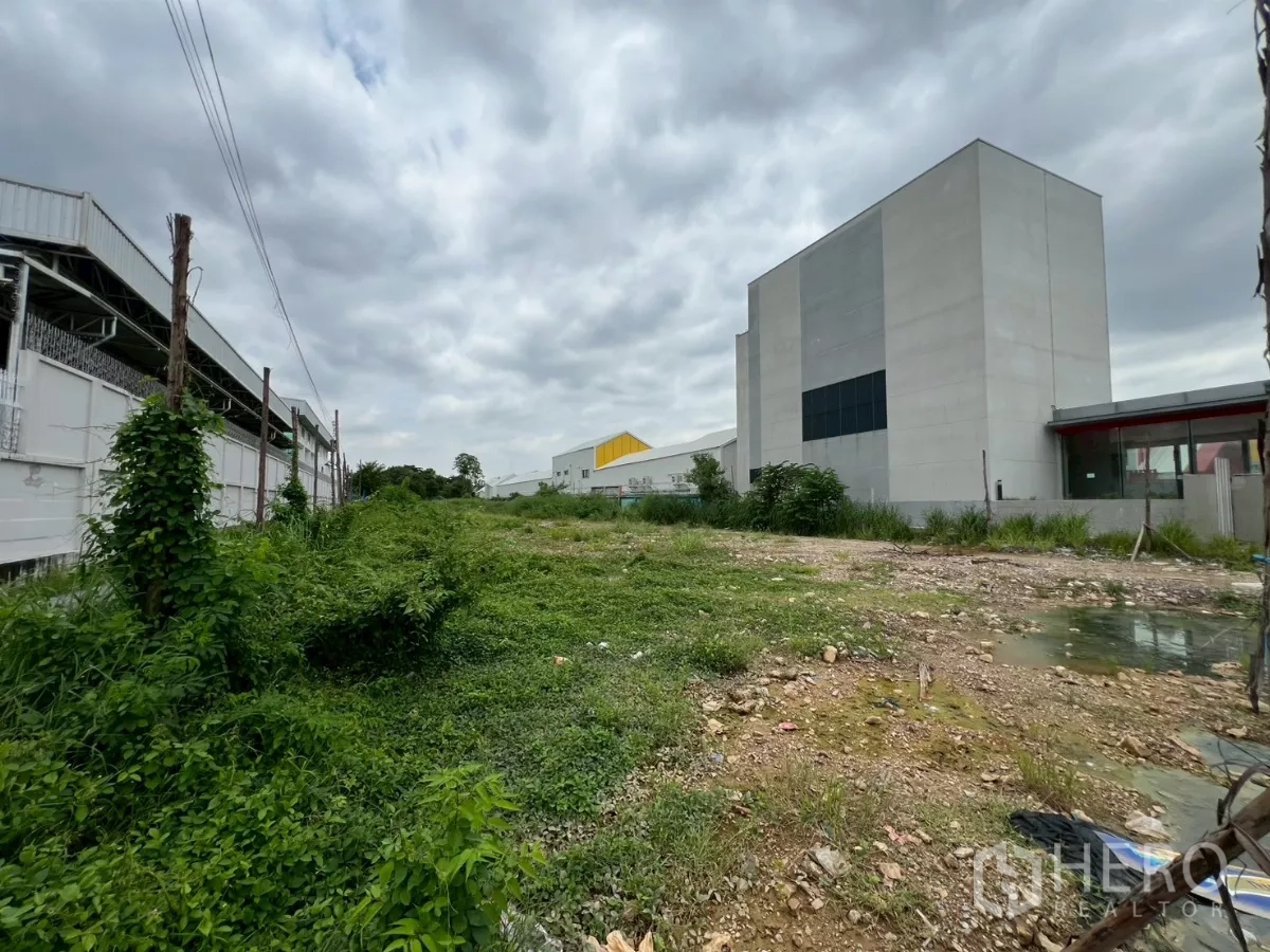 Land for sale - Undeveloped plot bordered by factory buildings and green groundcover in an industrial zone.