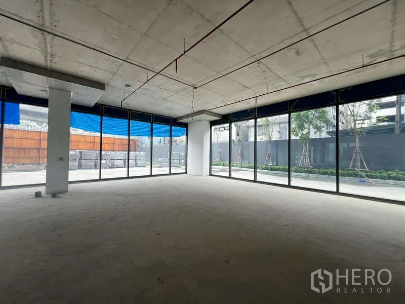 零售空间 出租 - 明亮的转角商铺毛坯空间，双面落地玻璃、挑高混凝土天花，面向景观步道，位于曼谷帕亚泰。