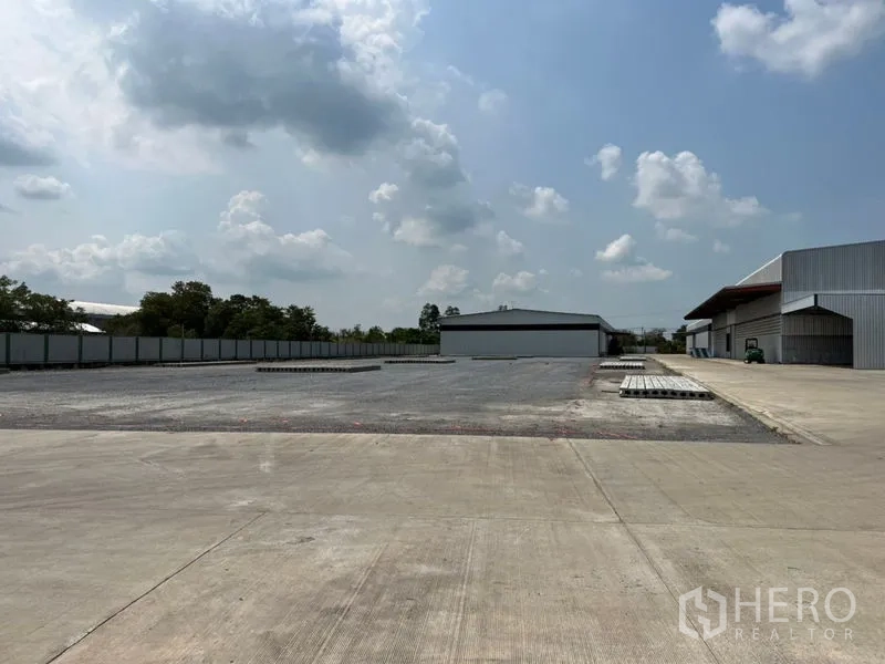 Warehouse for rent in Pathum Thani - Expansive concrete yard with perimeter fence and modern warehouse buildings under a cloudy sky.