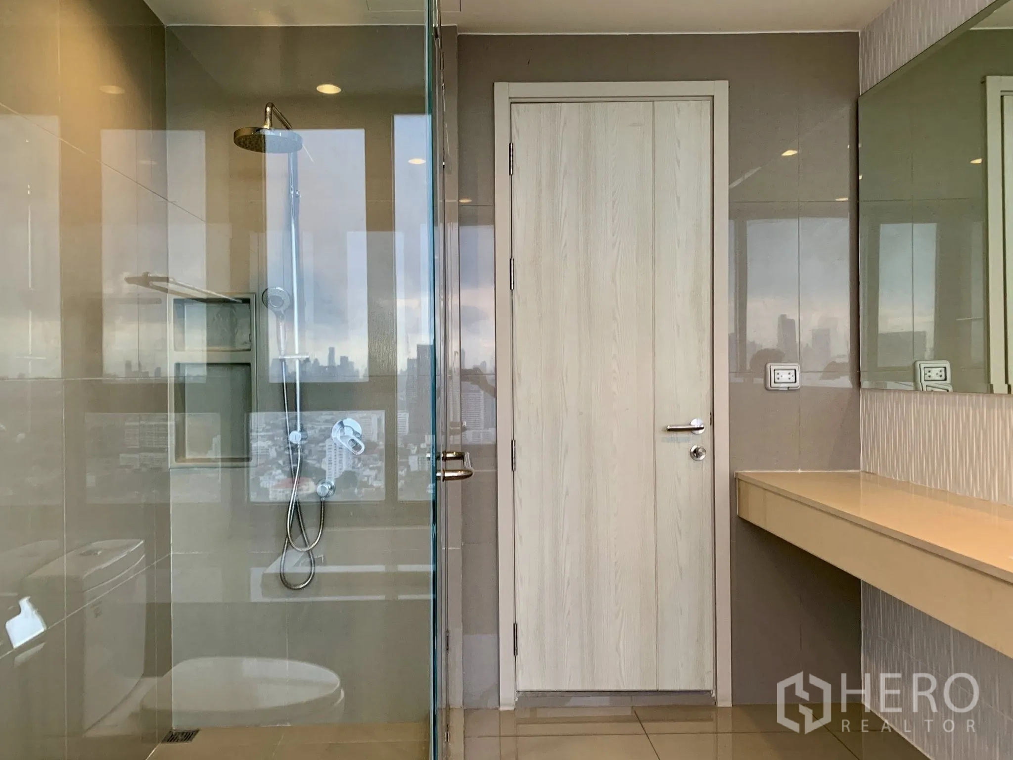 Condominium for rent in Khlong Toei, Bangkok - Modern bathroom with glass-enclosed shower, long vanity, and city reflections on the glass.