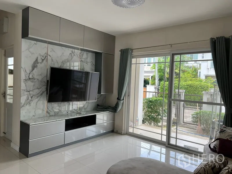 Detached House for rent in Bang Kapi, Bangkok - Living area with built-in TV cabinet and large windows with grey curtains.