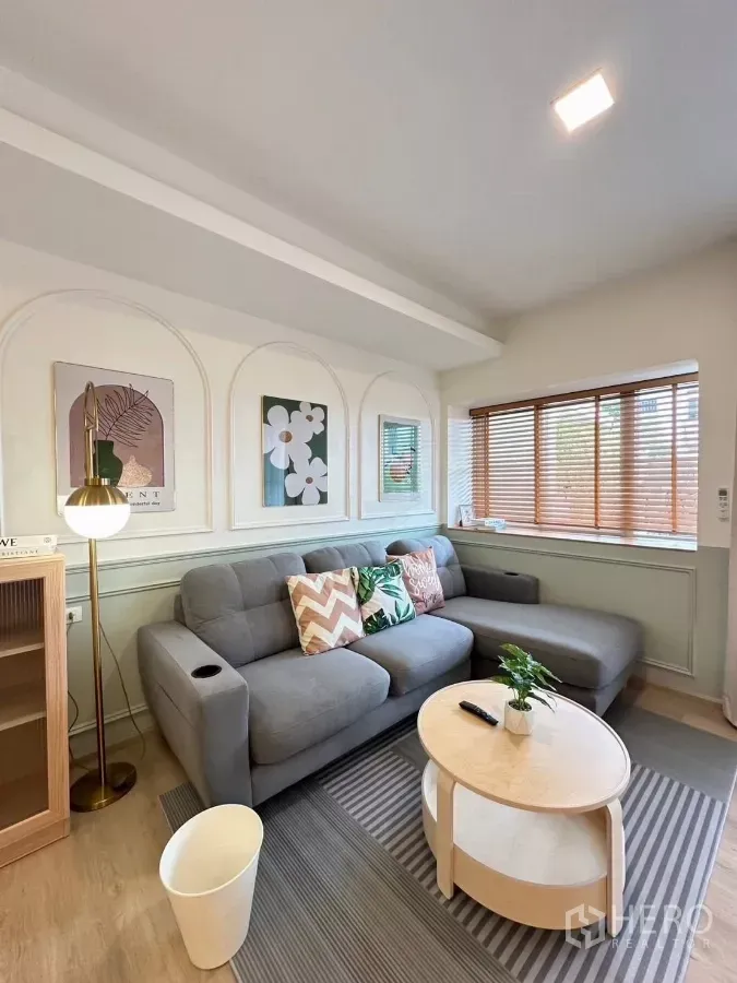 联排别墅 出租在Prawet, Bangkok - 温馨客厅，灰色L型沙发、圆形茶几、墙面装饰与百叶窗。