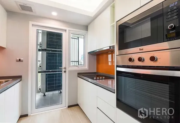 Apartment for rent in Huai Khwang, Bangkok - Modern closed kitchen with built-in oven and hob, white cabinetry, and glass door to a utility balcony.