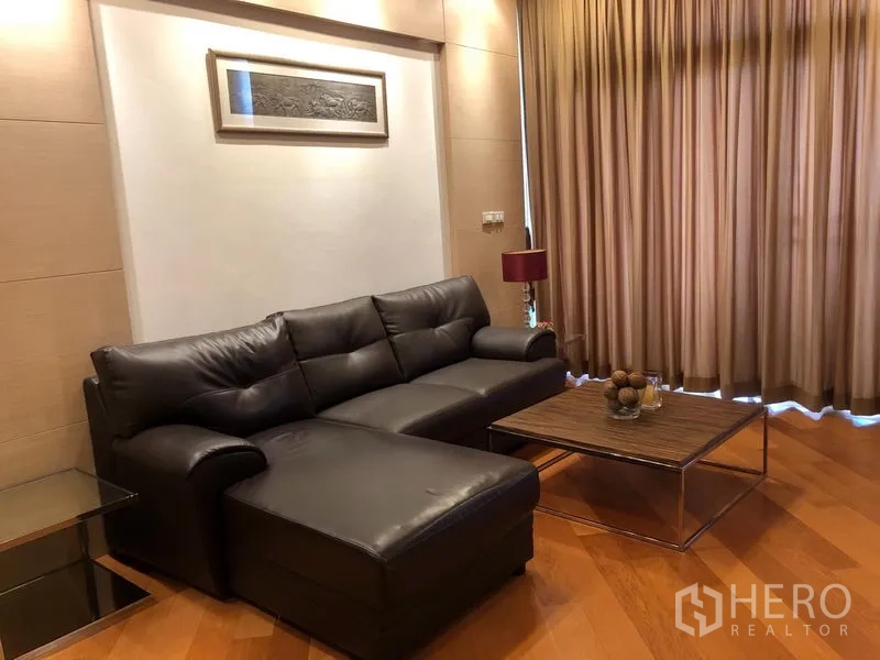 Condominium for rent in Bang Kho Laem, Bangkok - Cozy living room with leather sectional sofa, coffee table, and floor-to-ceiling curtains on warm wood flooring.