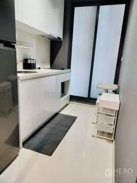 Condominium for rent in Khlong Toei, Bangkok - Compact galley kitchen with white cabinets, sink, hob and sliding glass door.