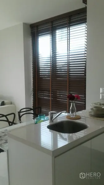 公寓 出租在Khlong Toei, Bangkok - 小巧厨房，白色岛台带水槽与花瓶，窗边木质百叶。