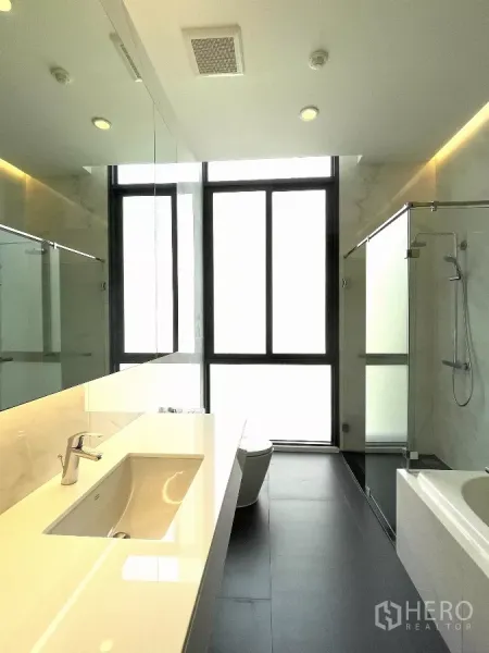 Condominium for rent in Bang Kapi, Bangkok - Modern bathroom with long vanity, glass-enclosed shower, bathtub and large frosted windows for natural light.