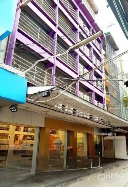 店屋 出租在Pathum Wan, Bangkok - 侧角街景：同一紫色商铺，玻璃店面与巷道上方招牌灯。