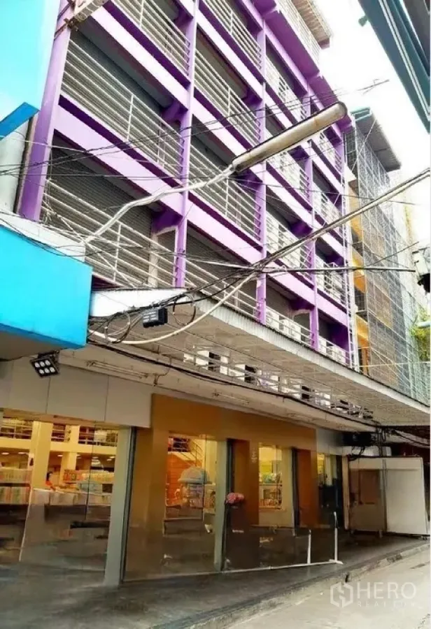 店屋 出租在Pathum Wan, Bangkok - 侧角街景：同一紫色商铺，玻璃店面与巷道上方招牌灯。