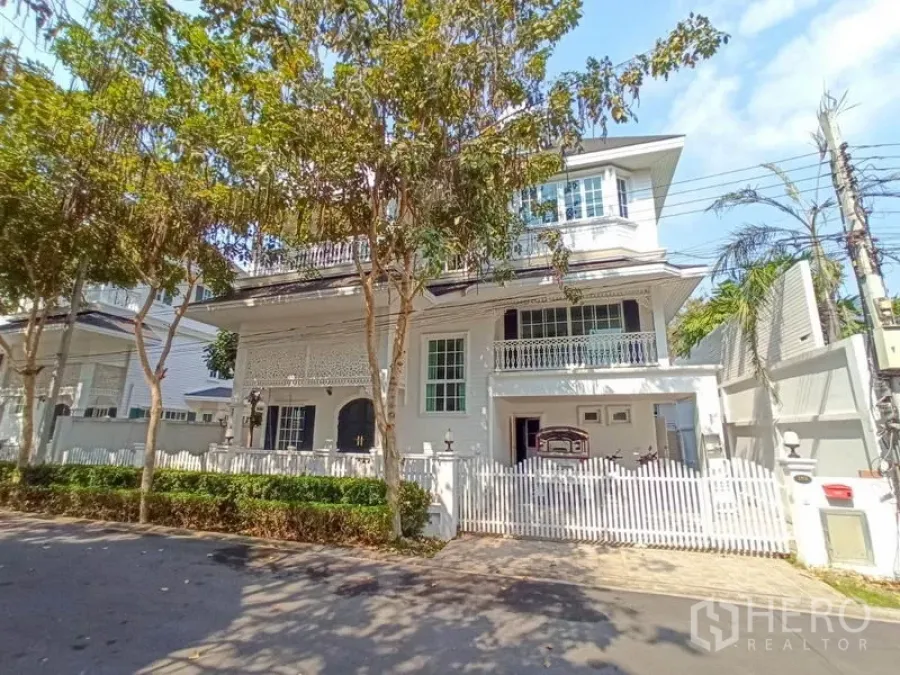 独立式住宅 出租在Mueang Samut Prakan, Samut Prakan - 白色独栋别墅外观，带阳台、白色栅栏和行道树，社区安静，位于沙没巴干。