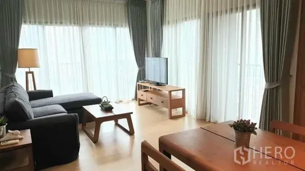 Condominium for rent or sale in Watthana, Bangkok - Corner living room with L‑shaped sofa, TV console and full‑height windows with sheer curtains.