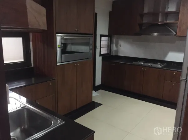 Detached House for rent in Khlong Toei, Bangkok - Separate kitchen with dark countertops, wood cabinets, built-in oven and gas hob.