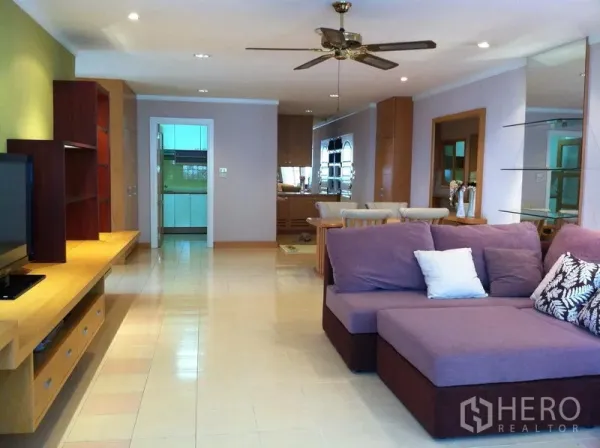 Condominium for rent in Watthana, Bangkok - Spacious Watthana Sukhumvit 39 condo living room with purple sectional sofa, built-in TV console, ceiling fan, and view to dining area and enclosed kitchen.