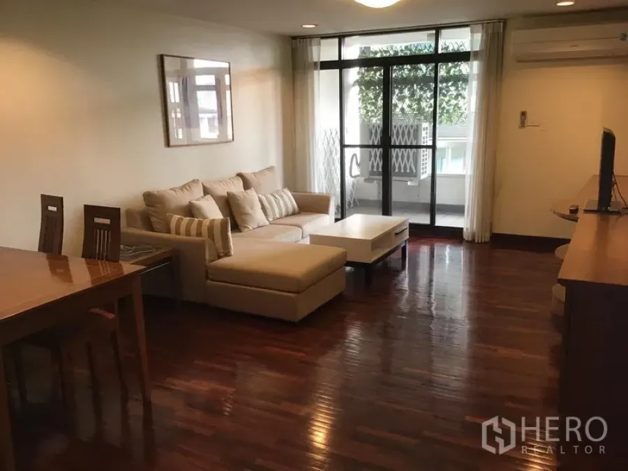 คอนโด ให้เช่าในWatthana, Bangkok - ห้องนั่งเล่นคอนโดโซนทองหล่อ พื้นไม้ โซฟาเข้ามุม และประตูกระจกสไลด์ออกสู่ระเบียงสีเขียว