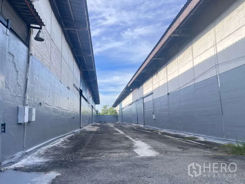 工厂 出租在Bang Pakong, Chachoengsao - 两栋灰色仓库之间的铺装装卸通道，金属屋顶与卷闸门。