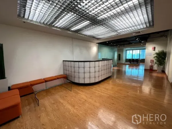 Office Space for rent in Khlong Toei, Bangkok - Spacious reception area with curved front desk and orange bench seating beneath a grid ceiling.