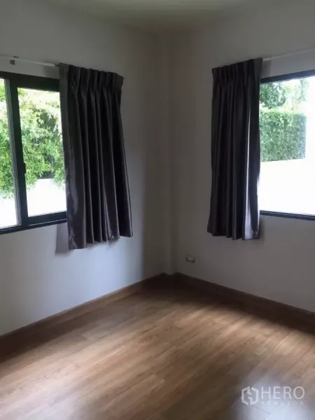 Detached House for rent in Samut Prakan - Sunlit bedroom with wood laminate flooring and two curtained windows facing greenery.