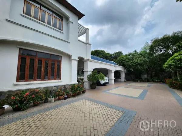 独立式住宅 出租在Prawet, Bangkok - 白色两层独栋别墅外立面，拱形车棚与宽阔铺砖车道，周边点缀花盆绿植。