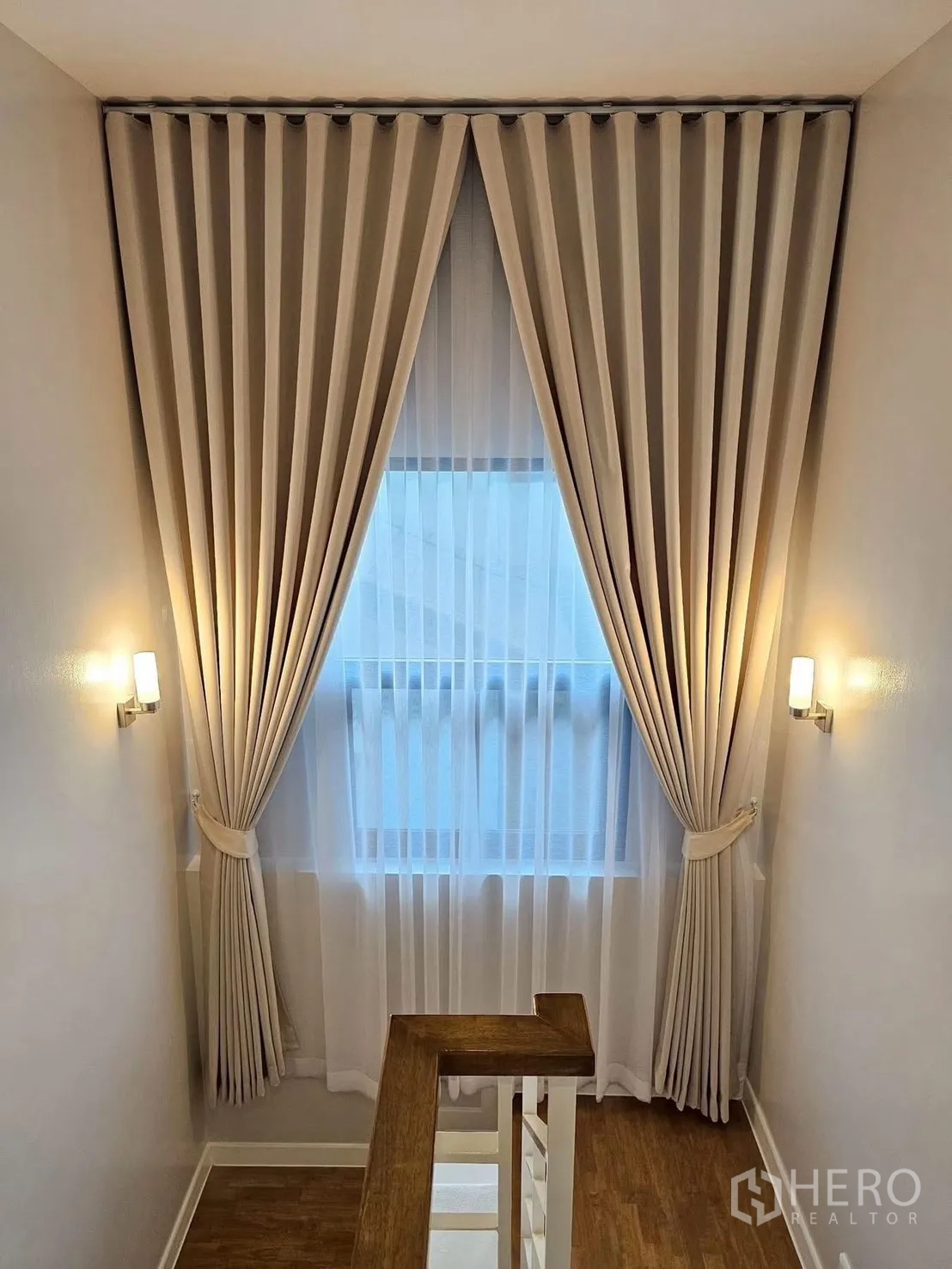 Detached House for sale in Bang Phli, Samut Prakan - Staircase landing with wooden handrail, tall window dressed in sheer and blackout curtains, and warm wall sconces.
