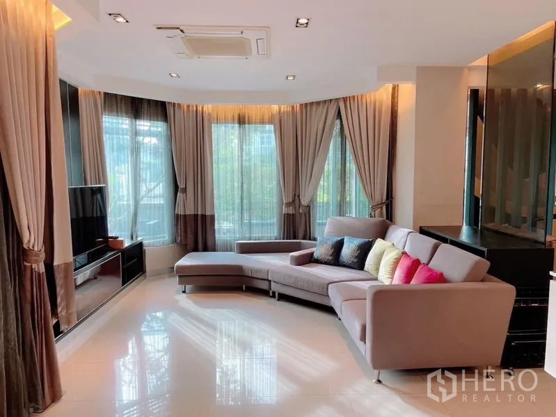 บ้าน ให้เช่าหรือขายในPrawet, Bangkok - มุมนั่งเล่นกระจกบานกว้าง ผ้าม่านทึบโปร่ง และโซฟารูปตัว L หันสู่ผนังทีวี.