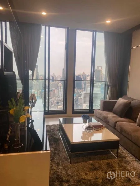 Condominium for sale in Pathum Wan, Bangkok - Living room with sofa and coffee table beside panoramic floor-to-ceiling windows.
