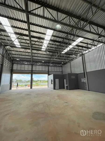 Warehouse for rent in Phra Samut Chedi, Samut Prakan - Open warehouse space with a large roller door facing the canal‑side rear yard and visible utility panels.