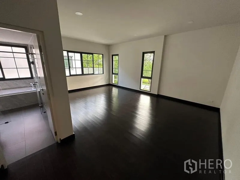 Detached House for rent or sale in Bang Phli, Samut Prakan - Expansive bedroom with dark wood flooring, corner windows and an ensuite bathroom visible through a doorway.
