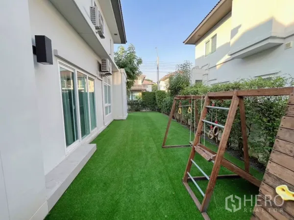 Detached House for rent in Samut Prakan - Side garden with artificial grass and a wooden children’s play set along the house exterior.