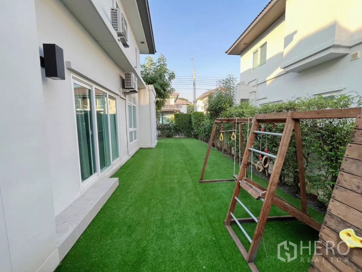 Detached House for rent in Samut Prakan - Side garden with artificial grass and a wooden children’s play set along the house exterior.