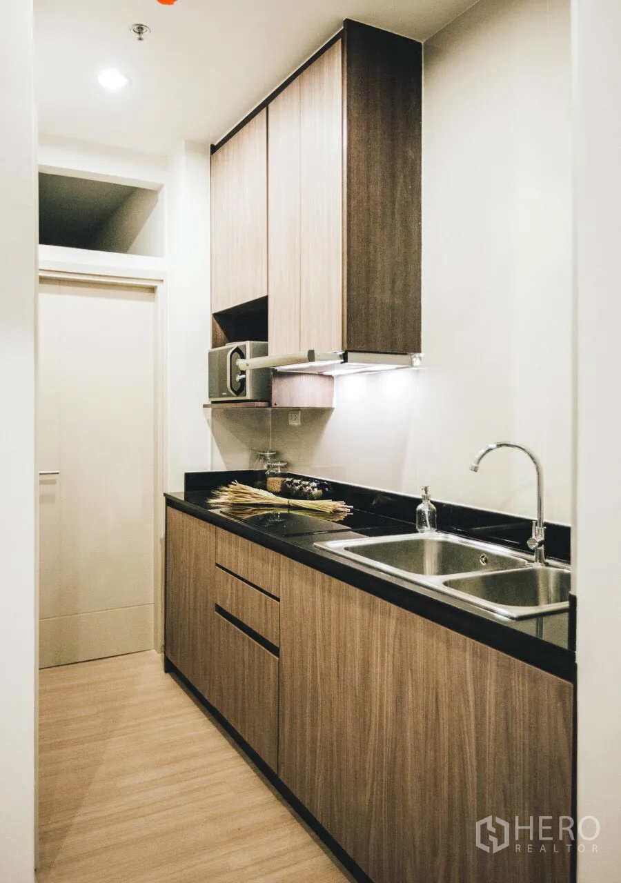 Condominium for rent in Huai Khwang, Bangkok - Compact galley kitchen with wood cabinetry, black stone countertop, cooktop, and double sink.