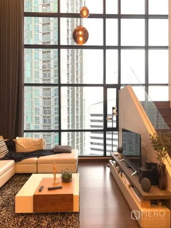 Condominium for rent or sale in Huai Khwang, Bangkok - Loft living room with oversized windows and staircase, city backdrop.