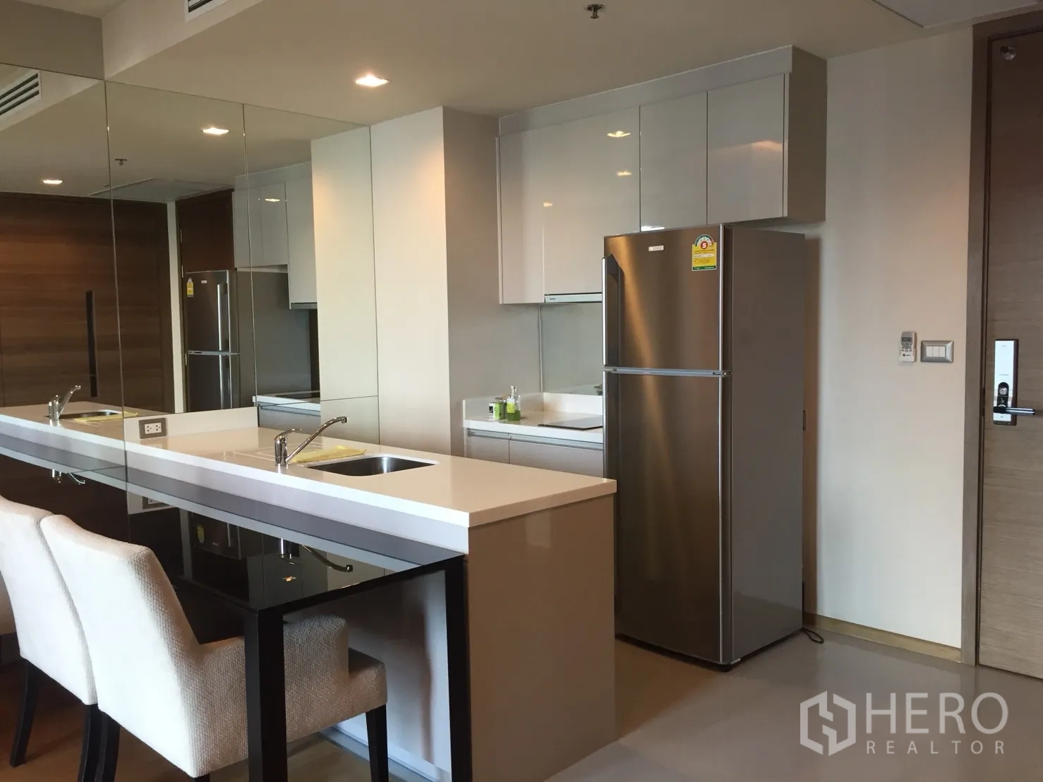 公寓 出租在Bang Rak, Bangkok - 现代厨房，白色橱柜、镜面墙、吧台以及不锈钢冰箱。