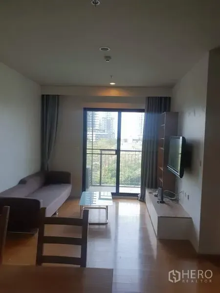 Condominium for rent in Watthana, Bangkok - Bright living room with sofa, TV wall and sliding door to a balcony overlooking greenery.