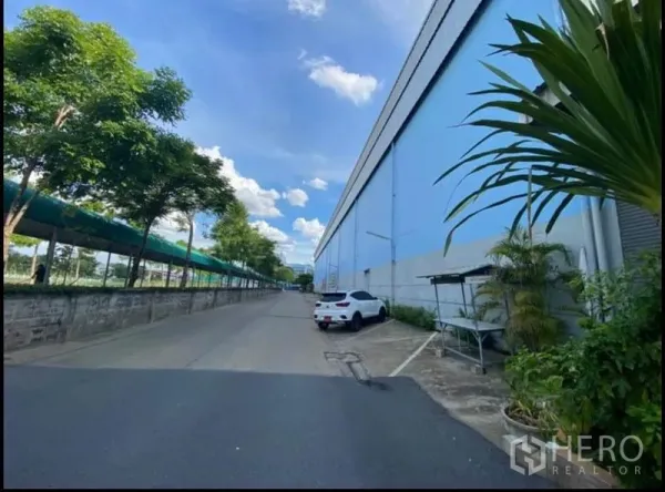 货栈 出租在Sai Mai, Bangkok - 仓库长侧立面，路边有车辆停放，绿树成荫的服务道路适合卡车通行。