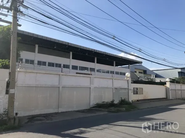 Factory for sale in Khlong Luang, Pathum Thani - Street view of a large gated factory compound with high roof and overhead power lines in Khlong Luang.