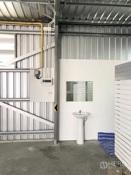 Warehouse for rent in Nonthaburi - Utility corner inside the warehouse featuring a white sink, mirror and exposed electrical piping against corrugated metal walls.