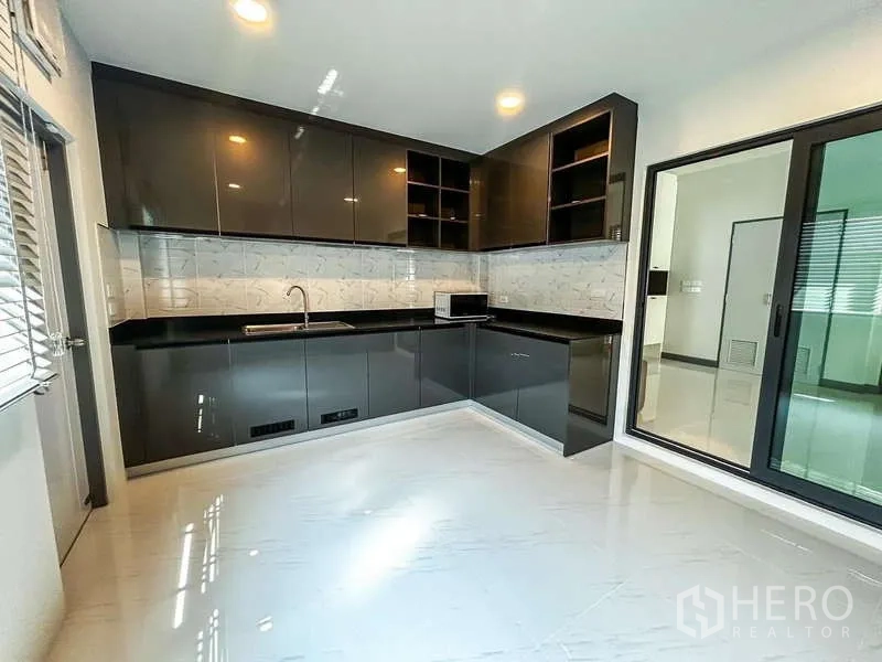 Detached House for rent in Bang Phli, Samut Prakan - L-shaped modern kitchen with glossy cabinets, stone-look backsplash, sink and sliding glass door.