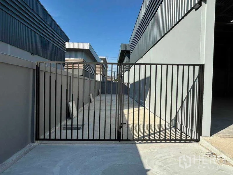 Factory for rent in Bang Bo, Samut Prakan - Secure metal gate opening to a side loading yard and service area between warehouse buildings.