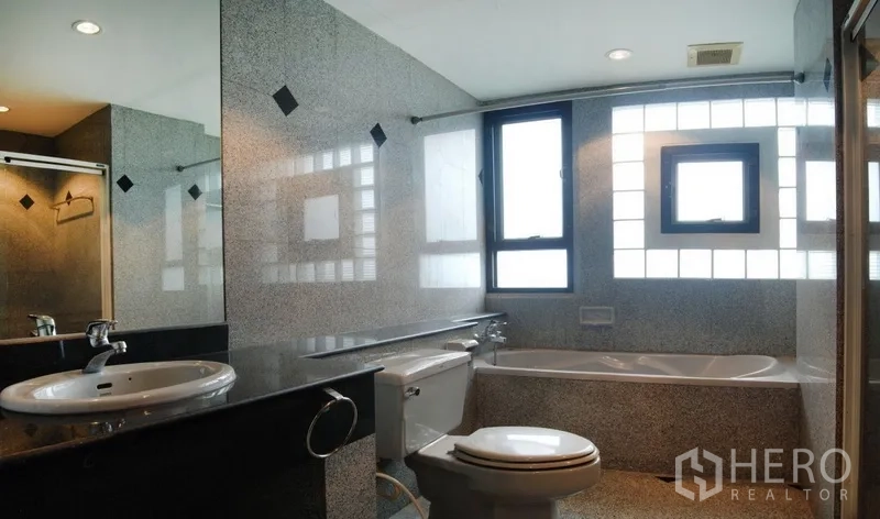 Condominium for rent in Watthana, Bangkok - Bathroom with bathtub beneath windows, long countertop and wall mirror in neutral tones.