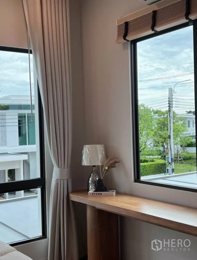 Detached House for rent in Samut Prakan - Upper-floor corner desk by two windows with garden outlook and a table lamp.