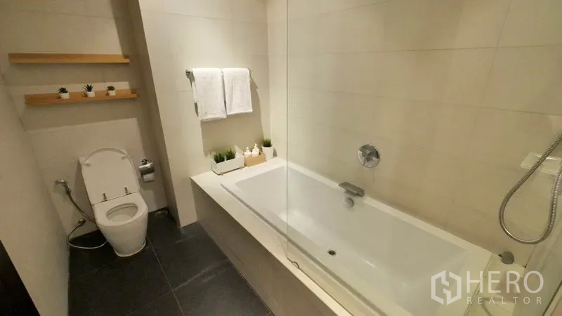 Condominium for rent or sale in Khlong Toei, Bangkok - Contemporary bathroom featuring a long bathtub with glass screen, handheld shower and toilet.
