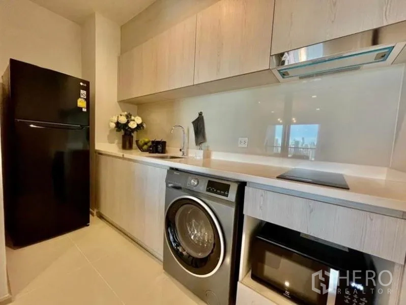 Condominium for rent in Pathum Wan, Bangkok - Modern galley kitchen with light wood cabinets, washer, cooktop, oven, and refrigerator.