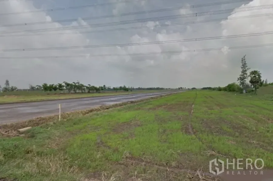 Land for sale in Lam Luk Ka, Pathum Thani - Wide green land plot beside a paved road with overhead power lines under a cloudy sky in Lam Luk Ka, Pathum Thani.
