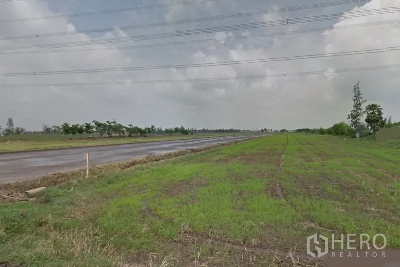 Land for sale in Lam Luk Ka, Pathum Thani - Wide green land plot beside a paved road with overhead power lines under a cloudy sky in Lam Luk Ka, Pathum Thani.